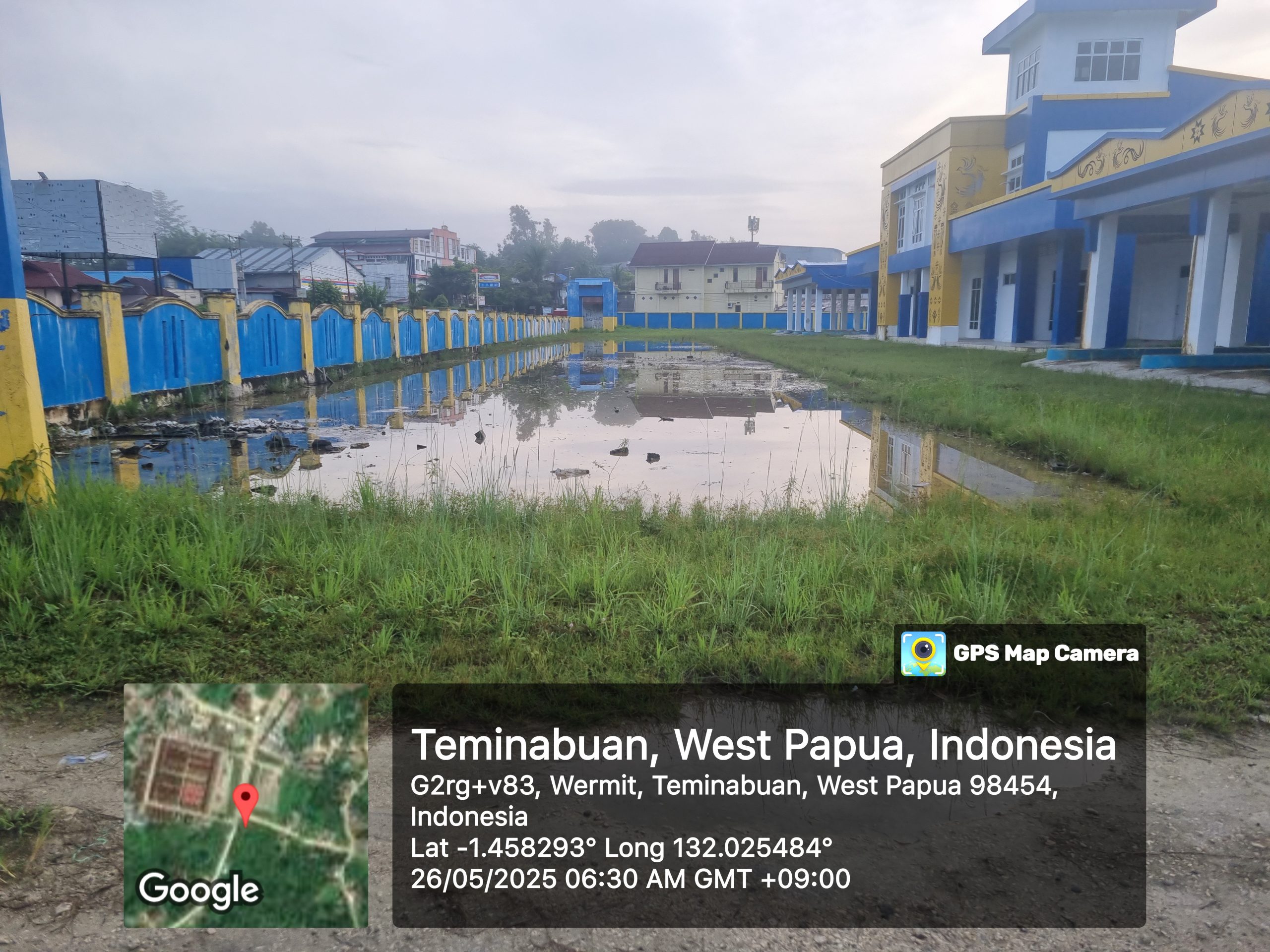 "Bangunan terminal berwarna kuning dan biru di Sorong Selatan tampak kosong dan tidak aktif, dengan halaman depan yang dipenuhi genangan air menyerupai kubangan. Rumput liar tumbuh subur di sekitarnya, dan pagar beton biru-kuning memanjang di sisi kiri gambar, mencerminkan kondisi area yang tidak terawat."