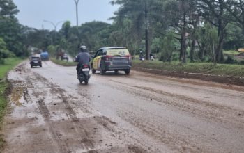 Alt text: Kondisi jalan di Kilometer 16, Kota Sorong, tampak berlumpur dan licin akibat tumpahan tanah dari truk proyek pembangunan gedung pemerintahan. Seorang pengendara motor terlihat berhati-hati melintasi jalan yang kotor dan berbahaya.